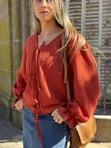 Blusa color terracota de muselina con escote en V y lazos para ajustar. Tiene mangas largas abullonadas con volados en los puños.