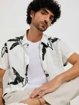 Camisa blanca de manga corta con estampado de hojas en negro y gris, cuello clásico y corte recto.