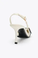 Zapato de tacón color crema con punta fina, tiras cruzadas en el empeine con hebilla dorada y tira trasera ajustable con hebilla.