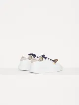 Zapatillas urbanas de cuero vacuno color blanco, con interior al tono y talonera en descarne color beige. Presentan doble cordón, uno azul y otro marrón, y dijes colgantes.