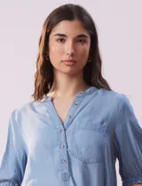 Blusa celeste de lyocell, con cuello a la base, escote en V y botones. Delantera con bolsillo y manga corta con terminación en punto smock.