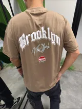 Remera color tostado de algodón con cuello redondo y corte amplio. Estampado en el frente con la palabra "Brooklyn" en tipografía gótica, la frase "New York" en cursiva y el logo "Bohemian Design".