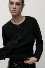 Chaqueta de punto negra con cuello redondo y manga larga. Presenta detalle de lazos con abalorios en el frente y cierre con botones a presión.