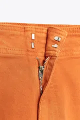 Pantalón de jean color naranja, de tiro alto y pierna ancha. Confeccionado en hilatura con mezcla de algodón. Presenta trabillas en la cintura, bolsillos de plastrón en la espalda y costura marcada en el delantero. Cierre frontal con cremallera, botón y gancho metálico.
