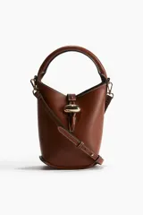 Cartera tipo balde color marrón con asa de mano rígida y correa bandolera ajustable y extraíble. Cierre con trabilla y adorno metálico dorado.