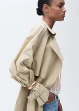 Abrigo tipo trench color beige, largo, con cuello de solapa, trabillas en los hombros y mangas largas abullonadas con trabillas en los puños. Incluye cinturón desmontable y bolsillos laterales.