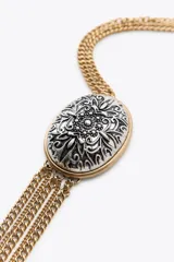 Collar largo con cadena dorada de eslabones metálicos. Colgante con medalla labrada en blanco y negro con flecos dorados. Cierre con mosquetón.