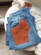 Pantalón de jean celeste con cintura elastizada, corte recto, bolsillos laterales y aplique de cuero marrón en el bolsillo delantero. Presenta botones metálicos en los laterales de la cintura y etiqueta de cuero en la parte trasera.