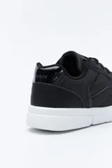 Zapatilla urbana blanca de mujer, con silueta moderna y líneas limpias. Presenta detalles en poliuretano liso y un panel plateado en el talón. La suela es de perfil recto y firme.