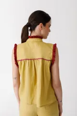 Blusa color mostaza sin mangas, con cuello alto y detalle de volados rojos en cuello, sisas y ruedo. Cierre frontal con botones.