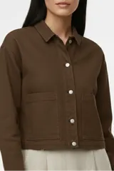 Chaqueta corta de algodón color marrón oscuro (Potting Soil), con cuello camisero, cierre frontal de botones plateados y dos bolsillos de parche en el frente.