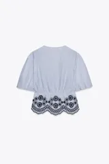 Blusa de rayas verticales azules y blancas, con escote en pico, mangas cortas abullonadas con elástico y bajo con bordados florales perforados. Cierre frontal con lazos.