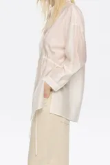 Camisa color crudo de corte ajustado en cintura, con cuello camisero, manga larga con puño y botón, cierre frontal con botonadura y túneles ajustables con tiras de tejido.