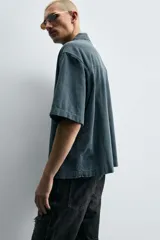 Camisa de denim de algodón, color beige claro, con corte relajado, cuello bowling y manga corta. Presenta cierre frontal con botones a presión y un pequeño bordado a tono en el pecho.