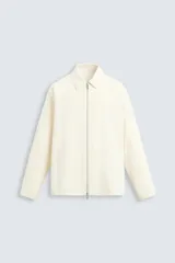 Sobrecamisa color crema de corte regular, confeccionada en tejido técnico ligero. Tiene cuello solapa, manga larga con puños abotonados, bajo ajustable con elástico en los laterales y cierre frontal con cremallera.