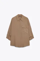 Camisa color marrón claro de cuello solapa y manga larga con trabilla y botón para remangar. Tiene un bolsillo de plastrón en el delantero, bajo asimétrico y cierre frontal con botones ocultos por solapa.