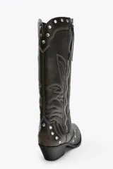Bota de caña alta color marrón oscuro con bordados en contraste y tachas plateadas. Cierre con cremallera frontal y tacón bloque bajo.