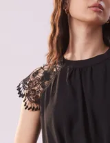 Blusa negra de crepe con apliques de crochet en los hombros. Tiene cuello redondo, mangas cortas y espalda con abertura ojal y botones.