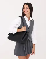 Cartera shopper de cuerina color negro con manijas superiores y charm a contratono. Tiene broche con botón para cerrar y cinturones laterales.
