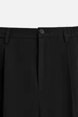 Pantalón gris de corte ancho con cintura elástica, pliegues frontales, bolsillos laterales y traseros de vivo. Incluye detalle de hebilla metálica en la trabilla y cierre frontal con cremallera y botón.