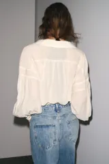 Camisa color crudo confeccionada en hilatura con mezcla de ramio, de corte holgado, con cuello solapa y escote pico. Tiene mangas largas con puños y detalle de tejido con pliegues.