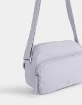 Bandolera gris claro de tela deportiva con bolsillo frontal, correa larga regulable, acceso con cierre superior y tag de goma.