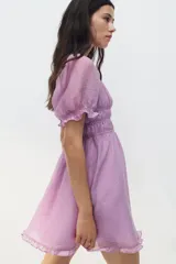 Vestido corto lila con escote cuadrado, mangas cortas abullonadas y fruncido elástico bajo el busto. Presenta volados en el escote, mangas y ruedo.