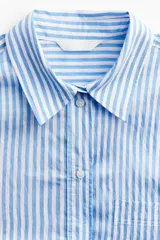 Camisa celeste y blanca a rayas verticales, de corte amplio, con cuello clásico, cierre frontal con botones, bolsillo de parche en el pecho, mangas largas con puños abotonados y bajo redondeado.