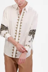 Camisa blanca de poplin de algodón con cuello clásico, cierre frontal con botones y mangas largas con puños abotonados. Presenta detalle de estampado de leopardo en blanco y negro en los puños y detalle de triángulos bordados en beige a lo largo del frente.
