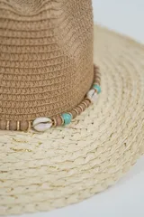 Sombrero de paja de ala ancha, con copa estilo fedora en color marrón y ala en color beige claro con hilos dorados entretejidos. Presenta una cinta decorativa con cuentas turquesas y un lazo de cordón fino atado alrededor de la base de la copa.