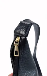 Cartera negra de cuero sintético con textura granulada, forma trapezoidal y asa de hombro ajustable con hebilla metálica.