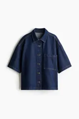 Camisa azul de denim, corte amplio, con cuello clásico, abotonadura frontal, manga corta, bolsillo de parche en el pecho y canesú en la espalda.
