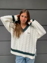 Sweater tejido de algodón color blanco, con cuello en V y ribetes a rayas verde oscuro y azul marino. Presenta un bordado pequeño en el pecho. Diseño de punto trenzado.
