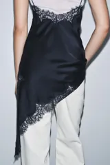 Vestido satinado negro con escote pico y tirantes finos. Presenta aplicación de encaje en el escote y en el bajo asimétrico, que incluye un tajo o caída pronunciada.