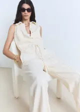 Mono largo color crudo, sin mangas, con cuello camisero, cierre frontal con botones, cintura ajustable con lazo y bolsillos delanteros y traseros. Diseño recto.