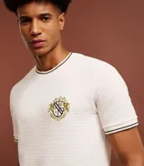 Remera de algodón y poliéster color crudo, con cuello redondo y manga corta. Presenta textura acanalada horizontal, estampa localizada con escudo y ribetes en contraste en cuello y mangas.