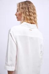 Camisa blanca de manga tres cuartos, con cuello camisero y corte holgado. Presenta un bolsillo de parche en el pecho izquierdo.