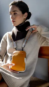 Cartera pequeña color mostaza con detalles en blanco, correa de tela con diseño de espiga en tonos marrones y grises, y bolsillo frontal con cierre.