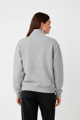 Buzo de algodón color beige claro (hielo) con cuello alto y cierre parcial frontal. Presenta un diseño holgado con franjas horizontales a la altura del pecho y texto estampado en color marrón oscuro.