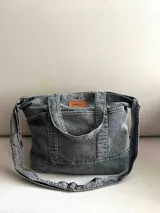 Bolso grande tipo totebag de jean azul, con asas de mano y asa larga para colgar. Se cierra mediante 1 botón de metal a presión y contiene 3 bolsillos exteriores.