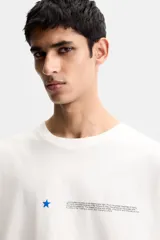 Camiseta blanca de corte holgado, cuello redondo y manga corta. Presenta un pequeño estampado de una estrella azul y un texto en negro en el frente.