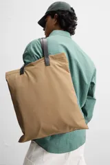 Bolso tipo shopper color beige, fabricado en tejido técnico con asas de mano de cuero negro. Tiene compartimento principal con cierre magnético y bolsillo interior con cremallera.
