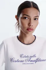 Camiseta blanca de algodón con cuello redondo y detalle de tricot a contraste en azul marino. Estampado frontal con texto "Estate sulla Costiera Amalfitana" en azul marino.