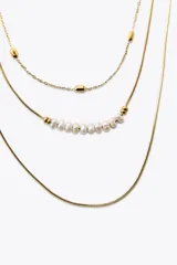 Set de tres collares dorados de diferentes largos. Uno es de eslabones metálicos con piezas circulares, otro es de cordón metálico con abalorios y perlas naturales, y el tercero es de cordón metálico liso.