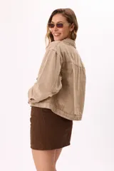 Campera de denim color beige, corte cropped, con cuello clásico, mangas largas abullonadas con puños abotonados, cierre frontal con botones metálicos y bolsillo de parche en el pecho.