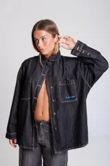 Camisa de denim azul oscuro, con cuello, bolsillos frontales con tapa y bordados en rojo en el bolsillo derecho. Presenta un calce holgado y mangas largas.