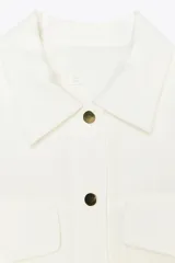 Camisa blanca de manga corta con cuello solapa, bolsillos frontales con tapeta, bajo con elástico y cierre frontal con botones a presión.