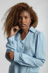 Camisa celeste de textura arrugada, con cuello camisero, escote en V y mangas largas abullonadas con puños abotonados.