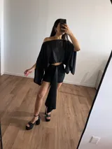 Conjunto negro brillante compuesto por un top corto con escote off-shoulder y mangas anchas, y una falda corta con lazo lateral.