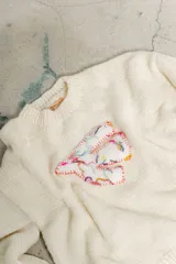 Sweater tejido color crudo con cuello redondo y parche bordado en el frente.